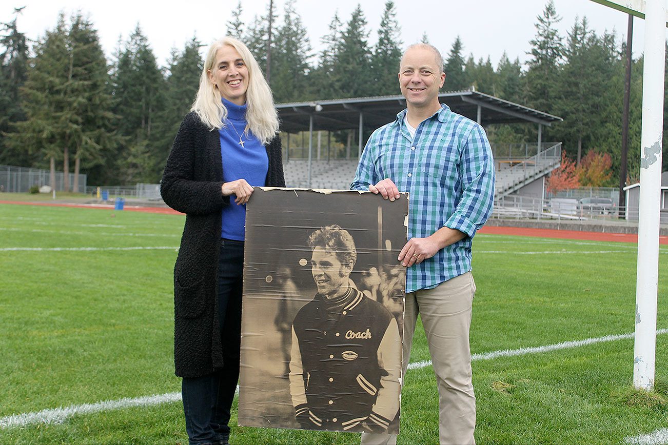 Evan Thompson / The Record Jim “Coach” Leierer’s daughter, Nanette Pierson (left) and community member Jon Chapman (right) stand on South Whidbey High School’s football field next to a picture of the deceased coach. The school board unanimously approved the stadium be renamed after longtime football coach Jim “Coach” Leierer on Wednesday night.