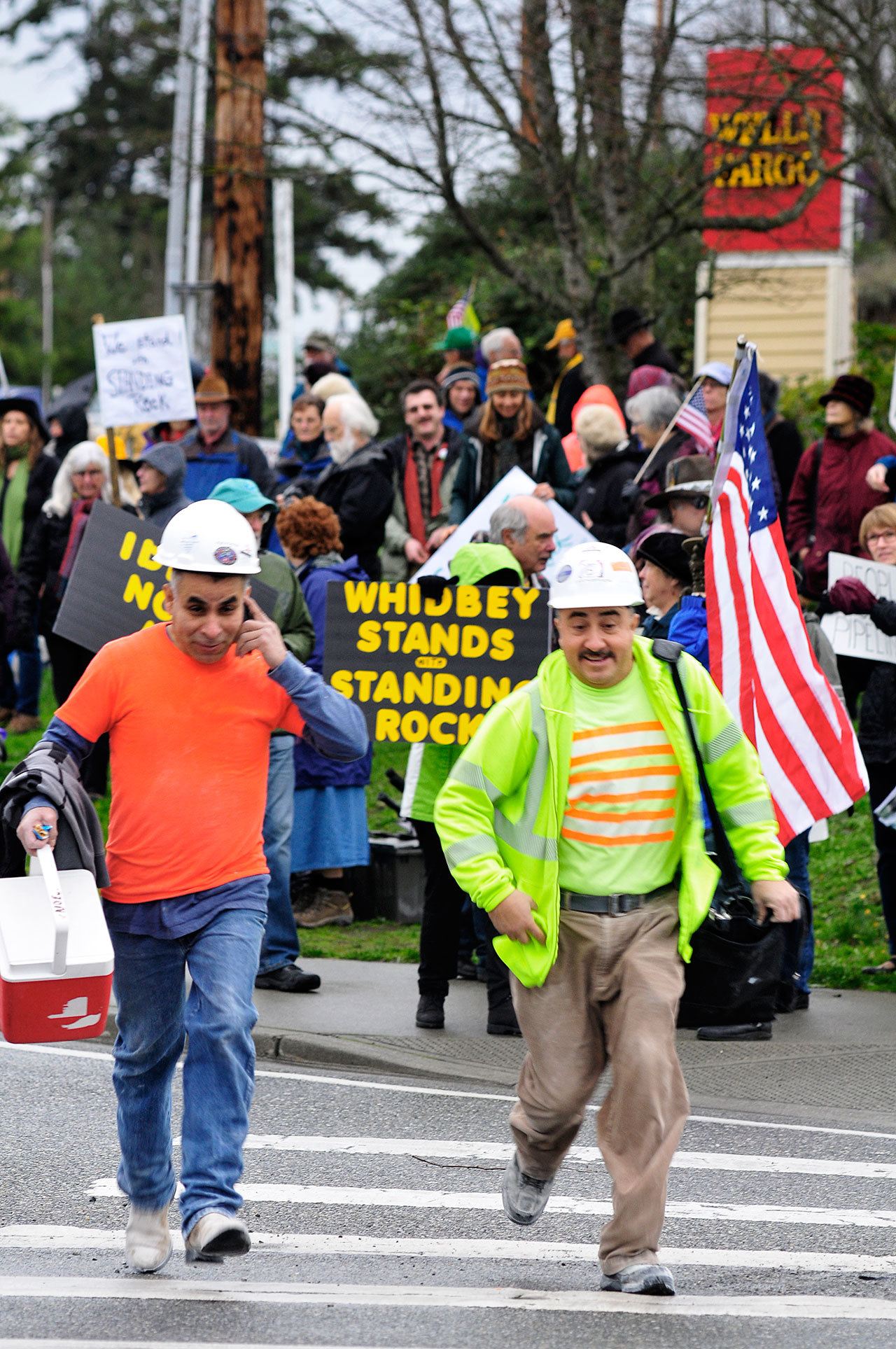 Photo by Michael B. Watkins/Whidbey News-Times                                Construction workers from WhidbeyHealth cross at State Highway 20 in Coupeville as protesters behind them protest the Dakota Access Pipeline stand outside Wells Fargo Bank in Coupeville last week. The group opposes the bank funding the pipeline project and the potential impact to the environment and native people’s land.