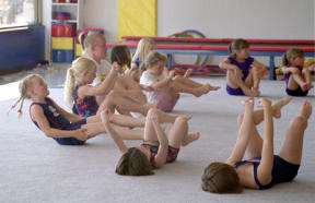 Girls in a beginning gymnastics class try to write their names in the air by circling their wrists and ankles. Coach Jennifer Cripps circles along with the class. The instructor/student ratio depends on the ages of students: in younger classes to ratio is only 7 to 1; in older classes it’s 9 to 1.