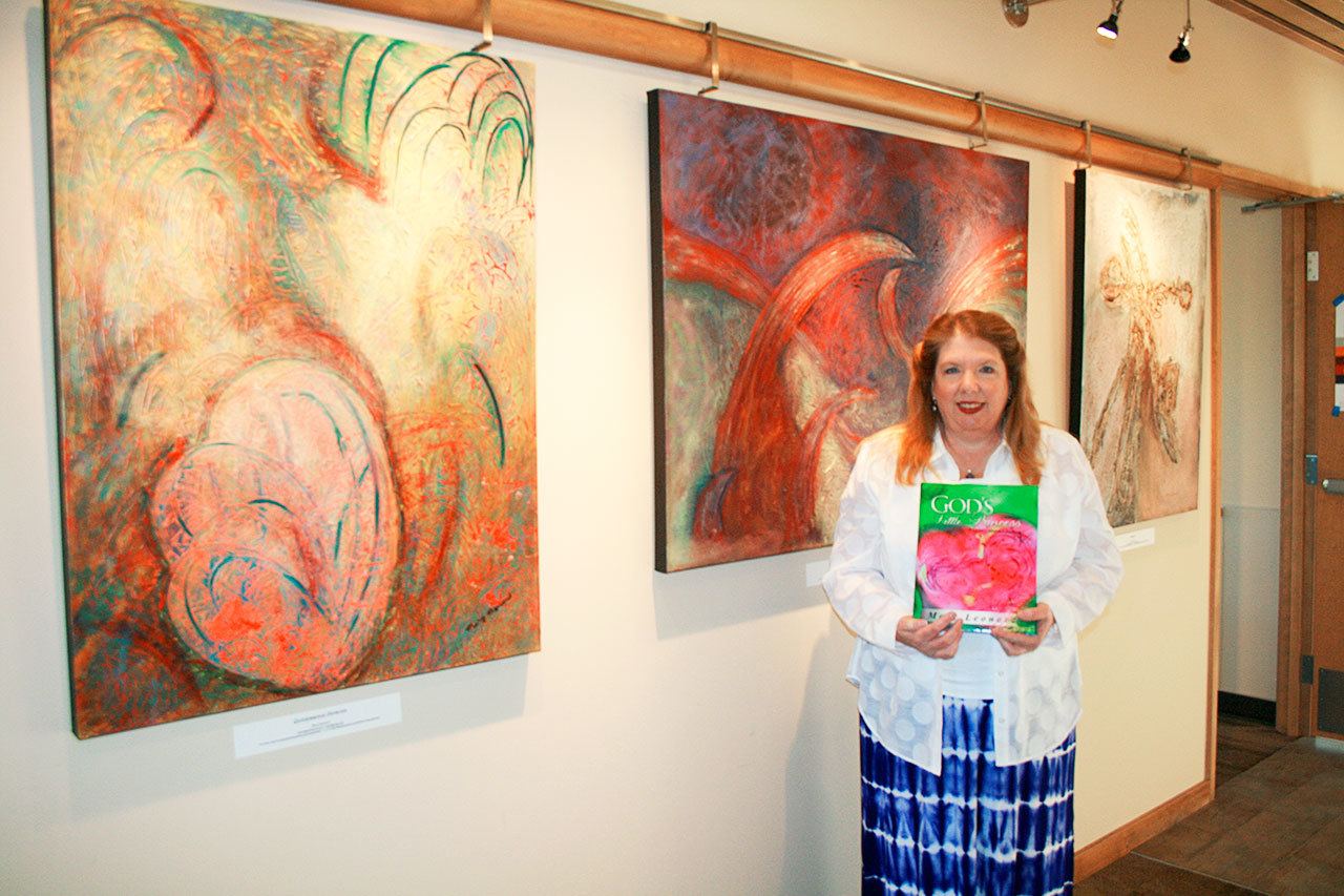 Mary Leonard, local artist and author, shows off her book, “God’s Little Princess; A Child’s Abstract Journey,” amid a display of her paintings at the Coupeville Library. She will be available for book signings at 11 a.m. Saturday, Feb. 25, at the Salty Mug in Coupeville. Photo by Dan Warn/Whidbey News-Times