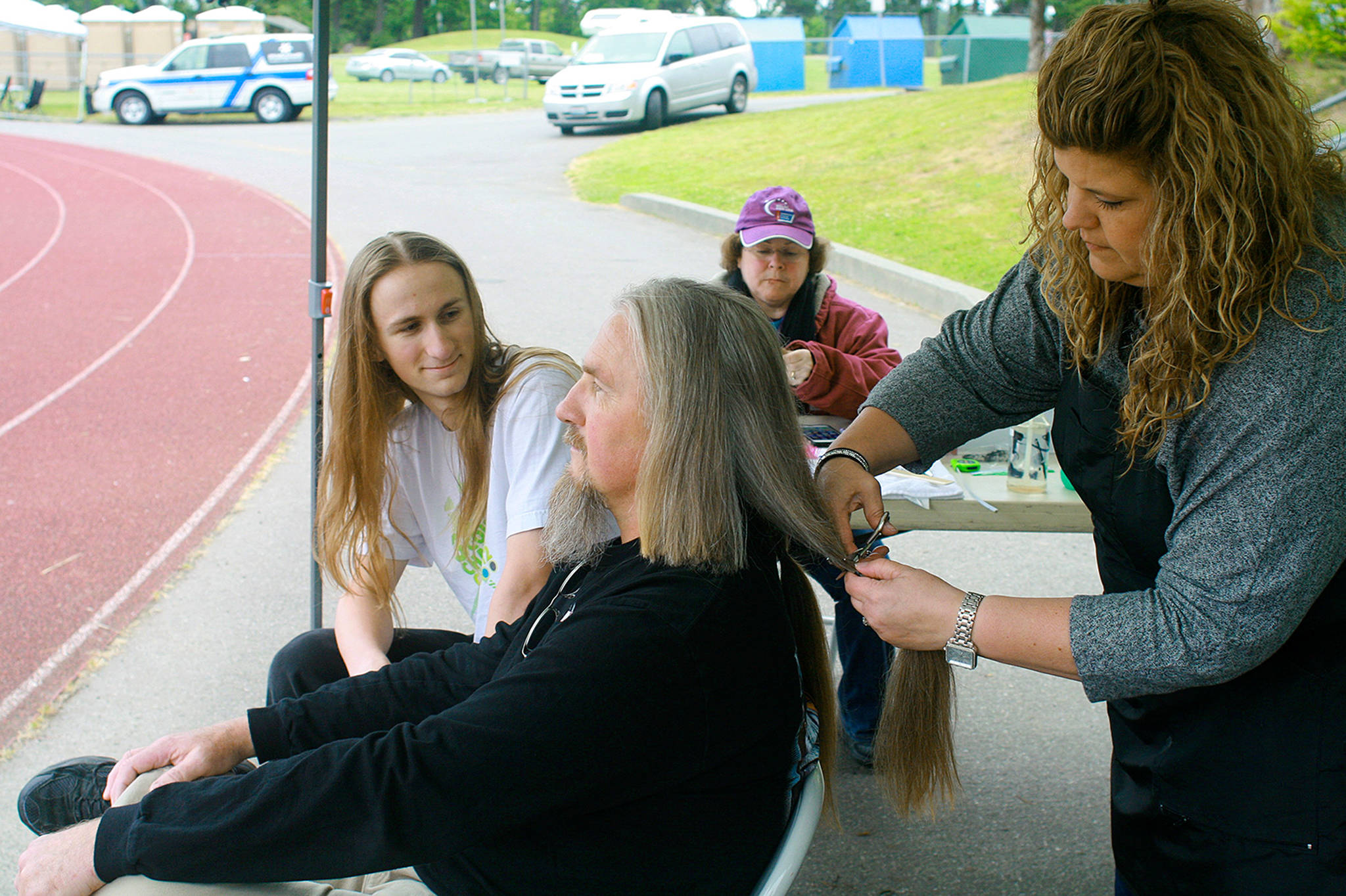 Les Richardson, center, gets two and a half years of hair growth clipped off by Oak Harbor resident Mollie Brodt at the Oak Harbor Relay for Life Saturday. Nathaniel Richardson looks on, awaiting his turn at the sheers. The Richardsons are donating their hair to Wigs For Kids to benefit cancer patients. Photo by Daniel Warn/Whidbey News-Times