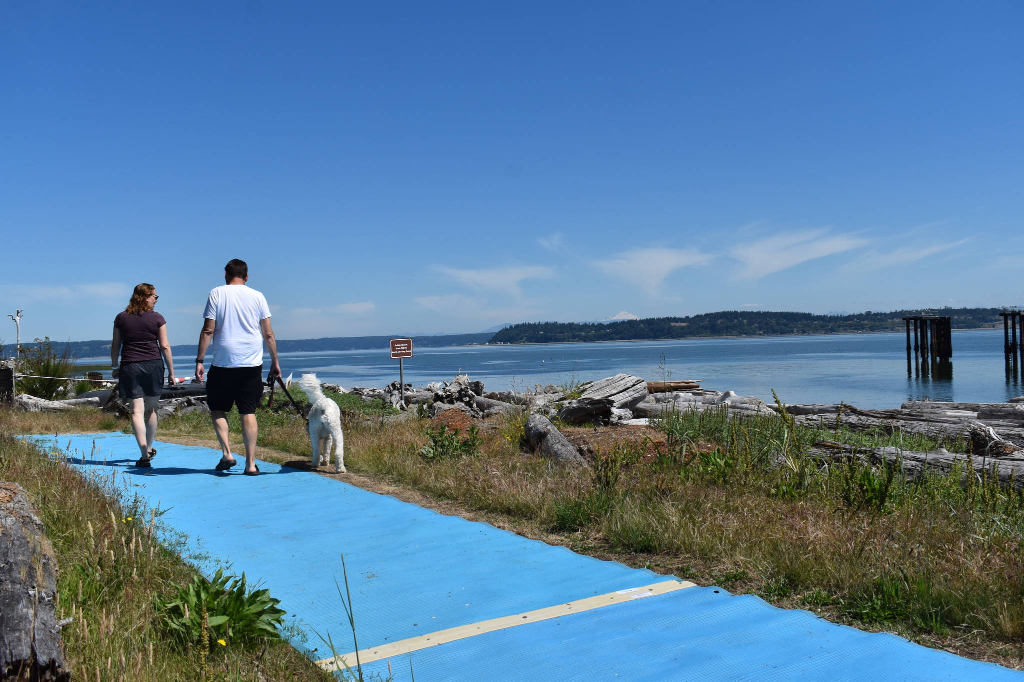 Photo by Emily Gilbert/South Whidbey Record A couple and their dog walk on the blue mats at Clinton Beach that volunteers from Island Beach Access installed for people with mobility issues. The mats cannot go all the way to the beach because of the amount of driftwood washed up by winter storms.