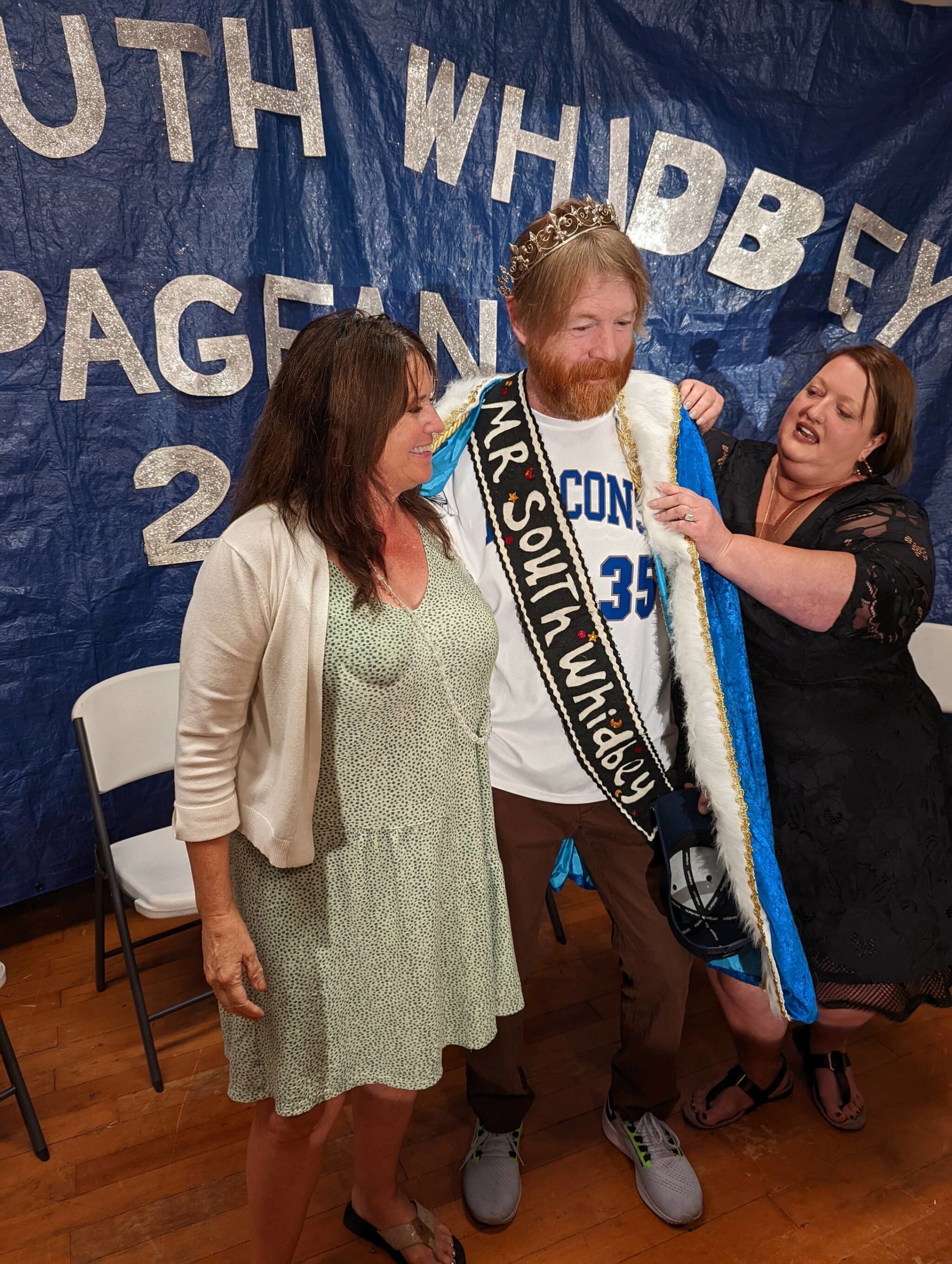 Photo by Sue Frause
Erik Stine, standing in for John LaVassar, was crowned Mr. South Whidbey this past weekend. Kristi Price, the board president for Friends of Friends Medical Support Fund, helped Stine into the Mr. South Whidbey robes while Gail, LaVassar’s wife, stood nearby.