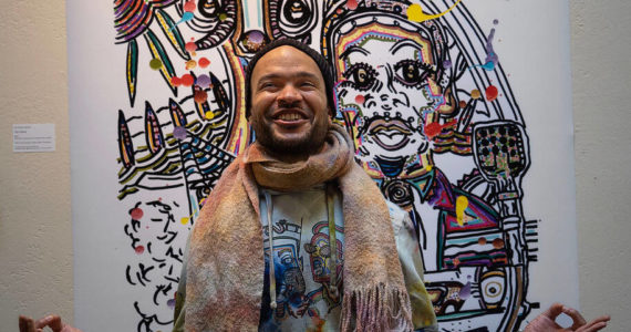 Photo by David Welton
Dressed in brightly tie-dyed clothing he made himself, Ian Joseph Jackson strikes a meditative pose in front of his artwork that includes Martin Luther King Jr. at the podium.