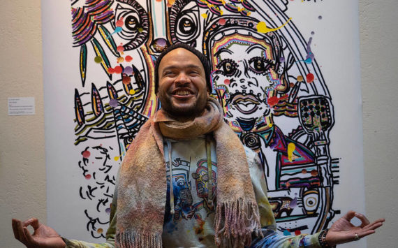 Photo by David Welton
Dressed in brightly tie-dyed clothing he made himself, Ian Joseph Jackson strikes a meditative pose in front of his artwork that includes Martin Luther King Jr. at the podium.