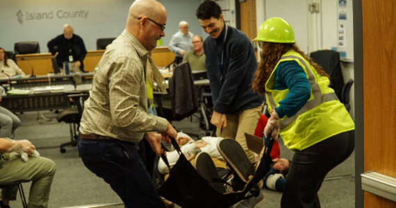 Island County Planning Manager Michael Jones (left), Health Director Shawn Morris (back) and Board Clerk Jennifer Roll (right) carry an actor during an earthquake demonstration at the commissioner's meeting on Wednesday. (Photo by Sam Fletcher)