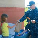 Rory Taylor, U.S. Navy petty officer second class, hands a free sack lunch, complete with a corn dog, to a neighborhood girl June 29 at Crescent Harbor Elementary, one of Oak Harbor Public Schools&rsquo; five free lunch sites. Photo by Daniel Warn/Whidbey News-Times