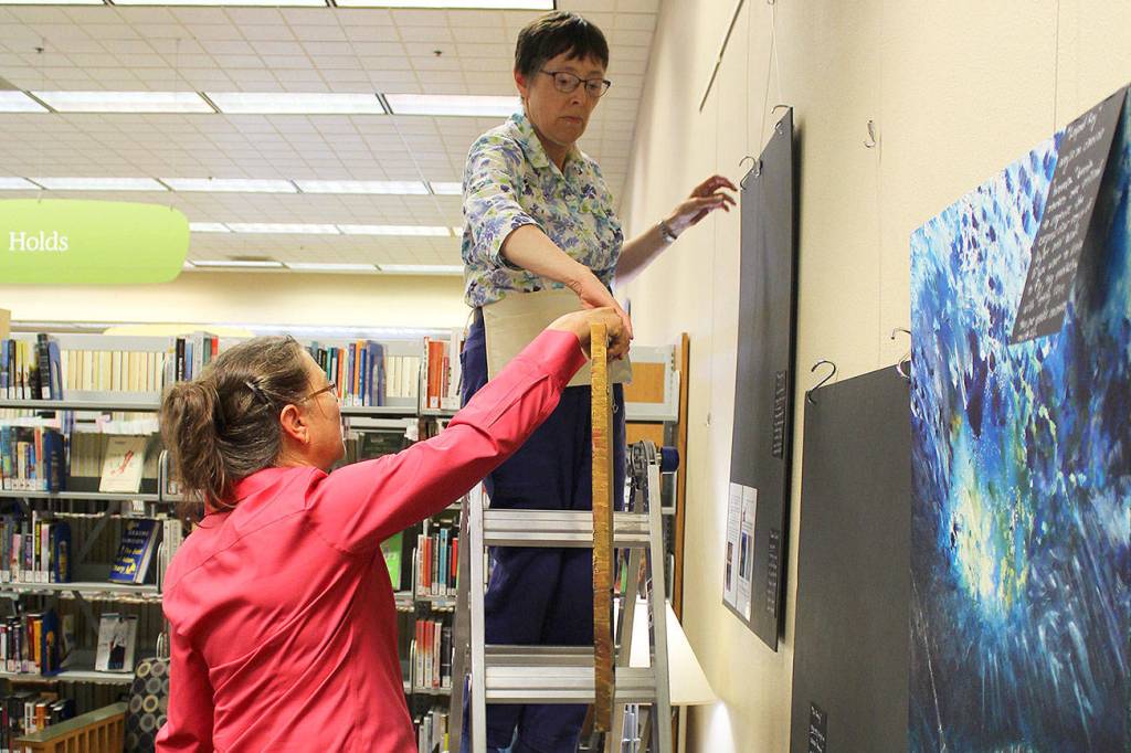 Tina Christiansen hangs her paintings at the Oak Harbor Library where she&rsquo;ll be the featured arist through August. She&rsquo;s assisted by librarian Nancy Luenn. Christiansen&rsquo;s art features landscapes of Alaska, sealife, birds and scenes from living near the sea.