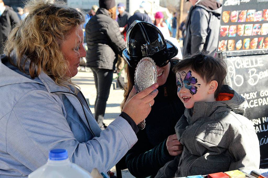 Sam Beaver, 3, takes in his reflection in the mirror held by face-painting artist Rochelle Walden, of Chelle Beautiful Face Painting. It has sparkles on it, Beaver said gleefully. (Photo by Maria Matson/Whidbey News -Times)