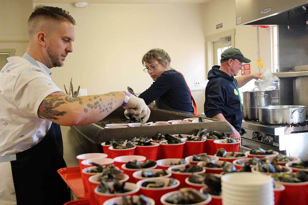 Kyle Johnson (left) helps get the cups of mussels ready for the mussel-eating competition. Rawle Jefferds of Penn Cove Shellfish monitors the stove while Laura Jefferds scoops up shellfish. Johnson was one of the chefs who demonstrated their cooking skills at the Coupeville Rec Hall. (Photo by Maria Matson/Whidbey News -Times)