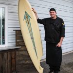 Steve Clarke poses by the surf board that welcomes customers to Slab City Sammies. Almost 20 year ago, Clarkes late pug Pugsley bit off a large chunk of the board, something Clarke and his wife decided to memorialize and pay homage to. (Photo by Luisa Loi)
