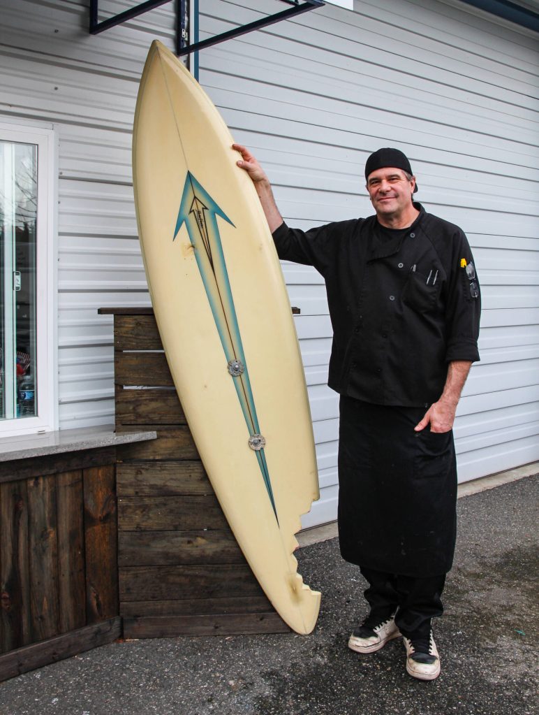 Steve Clarke poses by the surf board that welcomes customers to Slab City Sammies. Almost 20 year ago, Clarkes late pug Pugsley bit off a large chunk of the board, something Clarke and his wife decided to memorialize and pay homage to. (Photo by Luisa Loi)