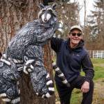 Pat McVay peeks out from behind the trunk of a mature Douglas fir that is adorned with his raccoon sculptures. (Photo by David Welton)
